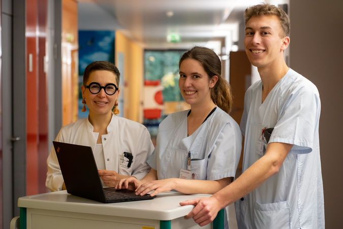 Eine weiblich gelesene Ärztin und zwei Pflegekräfte stehen an einem Stationswagen in einem Krankenhausflur.
