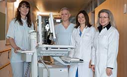 Vier weiblich gelesene Personen in weißen Kitteln und Kasacks stehen an einem Stationswagen in einem Krankenhaus.